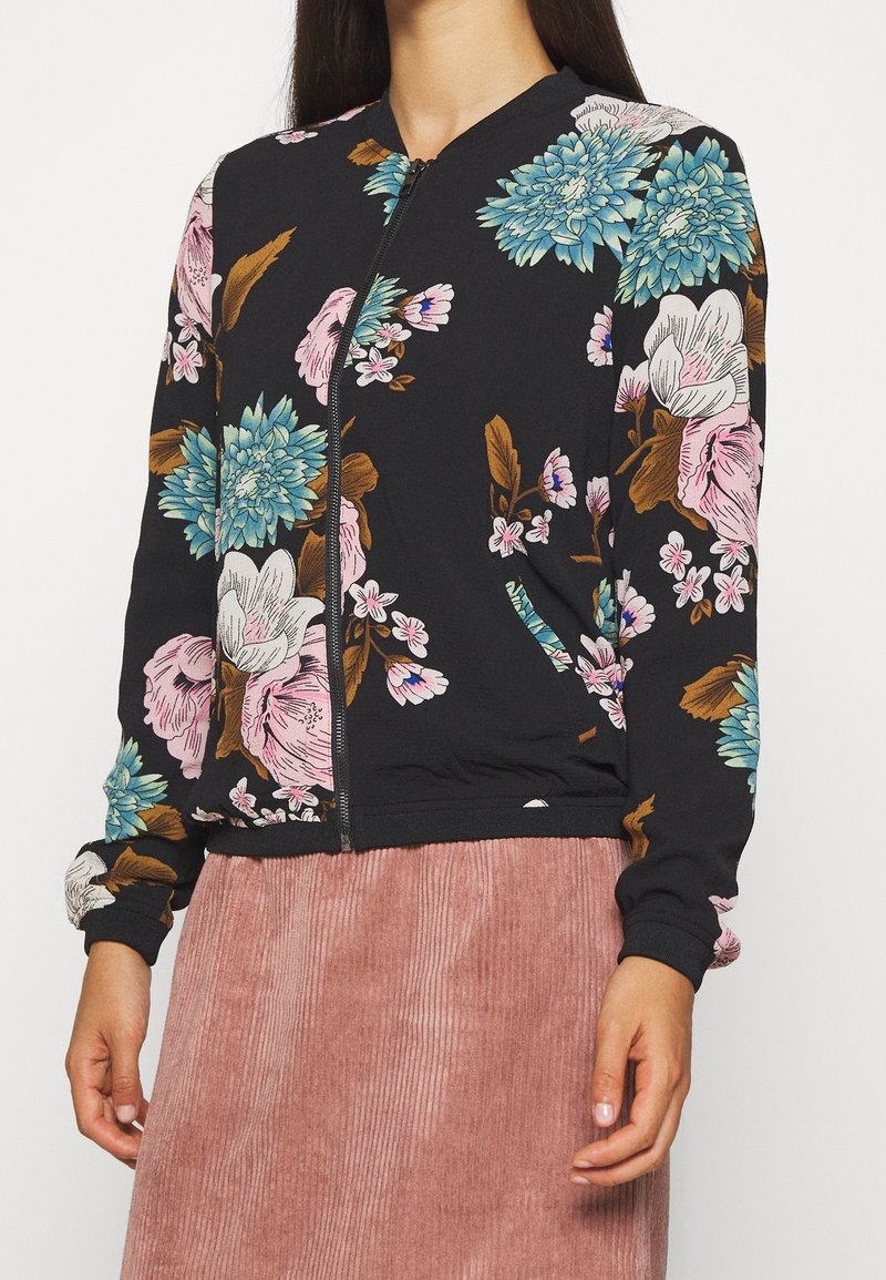 Giacca bomber floreale in tessuto nero con fiori blu, rosa e bianchi accompagnati da foglie verdi. Zip frontale e polsini e orlo a costine.