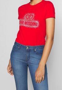 Rød t-shirt med korte ærmer og et skinnende sølvfarvet grafisk motiv af sammenflettede hjerter samt teksten "LOVE MOSCHINO." Sat sammen med blå jeans.