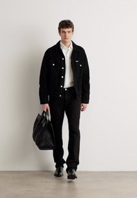 Veste en velours côtelé noire sur une chemise blanche, pantalon noir et chaussures noires. Portant un sac noir texturé. Design simple et contemporain.