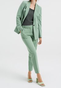 Kvinna klädd i en mintgrön kostym med skräddarsydda byxor och kavaj, kombinerad med en svart och vit randig topp samt mintgröna sandaler med öppen tå.