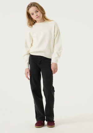 Giovane con i capelli lunghi che indossa un maglione bianco, jeans neri e scarpe rosse, in piedi di fronte su uno sfondo chiaro e semplice.