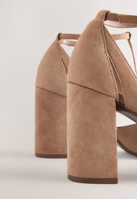 Beżowe sandały na wysokim obcasie z zamszu z masywnym obcasem, odkrytym palcem i paskowym wzorem. Teksturowana powierzchnia i jednolity kolor podkreślają styl.