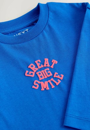 T-shirt en coton bleu avec un col rond en rib. Présente un texte rose en relief indiquant "GRAND SOURIRE" dans un design courbé. Texture lisse.