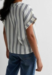 Blouse blanche à manches courtes avec des rayures verticales bleu marine. Portée avec un jean en denim bleu taille haute, affichant une coupe décontractée et des manches retroussées.