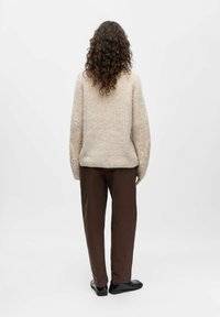 Bēša adīta džemperis ar fakturētu rakstu un dekoratīviem akcentiem uz piedurknēm, kombinēts ar brūnām biksēm un melniem apaviem. Skats no aizmugures.