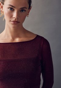 Dugih rukava bordo majica s kvadratnim izrezom na vratu, s teksturiranim dizajnom malih, istaknutih detalja po cijelom materijalu.