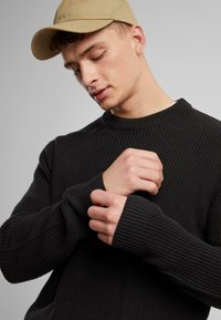Black ribbed sweater with a crew neck and long sleeves, displaying a textured knit. A tan cap is worn, partially obscuring the face.