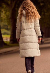 Un long manteau rembourré gris clair avec une capuche, présentant des sections matelassées. Porté par-dessus un pantalon côtelé foncé et des baskets blanches, vu de l'arrière.