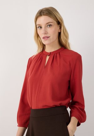 Vrouw met blond haar die een rode blouse met lange mouwen draagt met een klein met knoopjes gesloten sleutelgatdetail en donkere hoge taille broek tegen een effen achtergrond.