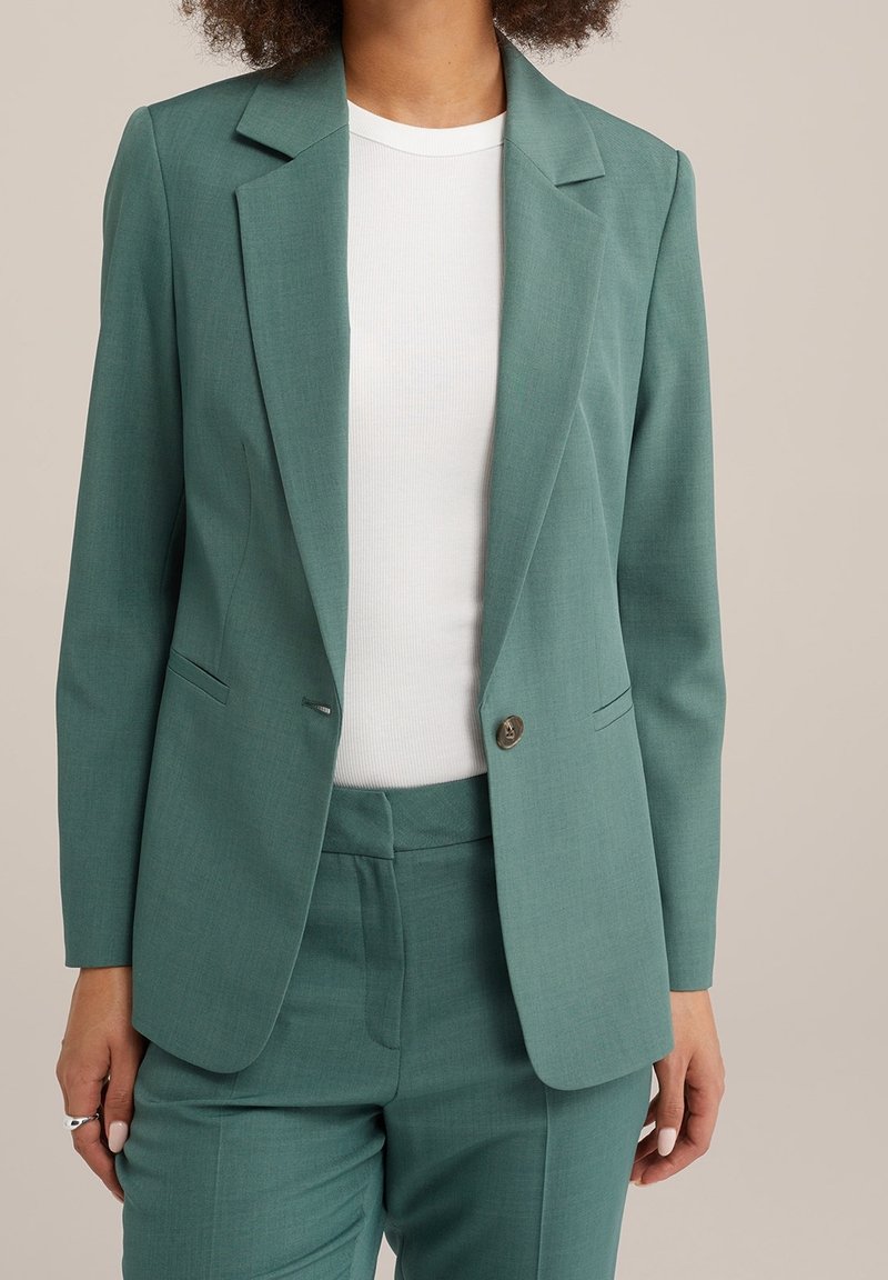 Femme portant un costume vert amande sur mesure avec une veste à un bouton et un pantalon assorti, sur une chemise blanche à côtes, debout devant un fond beige.