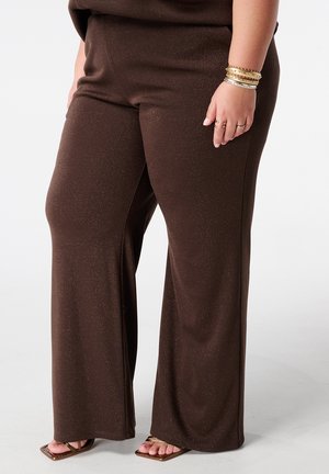 Vrouw die bruine broek met wijde pijpen, gouden armbanden, ringen en bruine sandalen met vierkante neus draagt, staand tegen een witte achtergrond.