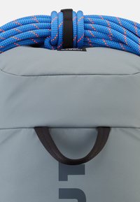 Grå ryggsäck med en strukturerad yta, med ett svart handtag och ett blått rep med orange detaljer fäst överst.