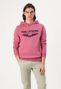 Różowa bluza z kapturem i kieszenią z przodu. Zawiera czarny nadruk logo z skrzydłami. Połączona z jasnymi spodniami.