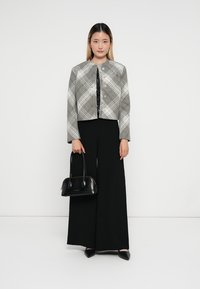 Giacca cropped a quadri grigia e bianca con colletto rotondo, chiusura con bottoni e maniche lunghe. Il modello indossa pantaloni larghi neri e tiene in mano una borsa nera.
