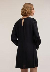 Vestito nero con maniche lunghe a sbuffo, caratterizzato da un'apertura a goccia sulla schiena e polsini decorati. Realizzato in un tessuto morbido e fluente.