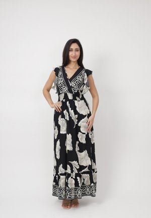 Femme debout portant une robe maxi noire et blanche à motif floral avec des manches à volants et un ourlet décoré, posant devant un fond blanc uni.