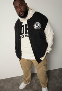 Chaqueta varsity negra y crema con un logo, combinada con una sudadera crema, pantalones cargo color beige y zapatillas blancas.