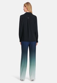 Schwarze Langarmbluse mit Kragen, mit einer Faltenlegung und einem goldenen Akzent. Kombiniert mit weiten Hosen in Blauverlauf, die zu Weiß übergehen.