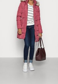 Abrigo de plumas rosa con capucha de pelo, camiseta a rayas blanco y negro, jeans azules y zapatillas blancas. Sosteniendo un bolso burdeos.