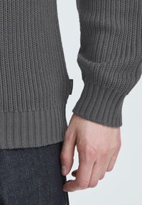 Maglione grigio a coste con superficie testurizzata. Presenta un polsino e un orlo aderenti. Piccola etichetta nera visibile sul lato vicino al fondo.