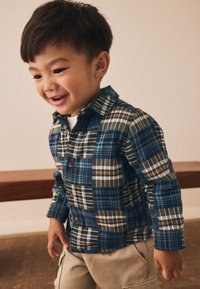 Blå og grøn ternet skjorte med knapper og forskellige lapper, blødt stof og brune knapper, båret af et barn med lyse bukser.