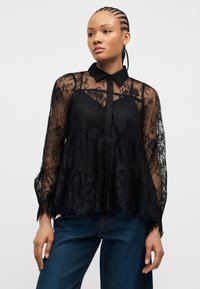 Γυναίκα με πλεγμένα μαλλιά φοράει μια διαφανή μαύρη δαντελωτή μπλούζα με λουλουδένια σχέδια και σκούρο μπλε τζιν, στέκεται μπροστά σε απλό φόντο.