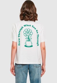 Hvid bomulds t-shirt med et grønt grafik af en smilende blomst og teksten "Vokse gennem det, du går igennem" på ryggen.