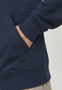 Dunkelblaues Sweatshirt mit strukturierten Rautenmustern im oberen Bereich, gerippten Bündchen und einer Seitentasche mit kontrastierenden Nähten.