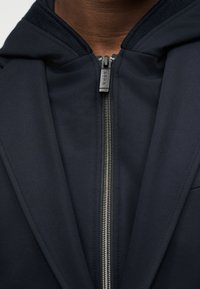 Κοντινό πλάνο ενός ατόμου που φοράει σκούρο μπλε μπουφάν με εμφανή ασημένια φερμουάρ και τσάκιση φερμουάρ με τη μάρκα "JOOP!"