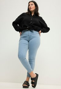 Camisa negra de botones con un nudo en la cintura, jeans ajustados con un patrón de color azul claro y sandalias negras con dos tiras anchas.