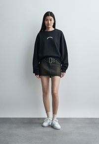 Νεαρή γυναίκα στέκεται, φορώντας μαύρο φούτερ, ξεθωριασμένη μαύρη μίνι φούστα με φαρδιά ζώνη, λευκές κάλτσες και λευκά αθλητικά παπούτσια σε απλό φόντο.