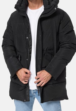 Homme portant une doudoune noire sur un t-shirt blanc et un jean bleu, ajustant la fermeture éclair de la veste avec ses deux mains.