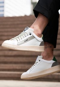 White sneakers with a mesh body, green and grey accents, and beige rubber soles. Features textured panels and green heel tab.