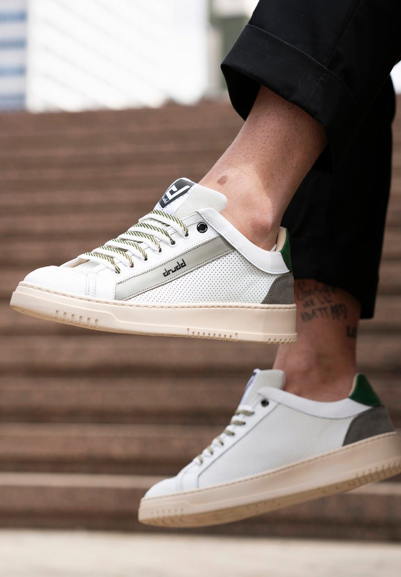 White sneakers with a mesh body, green and grey accents, and beige rubber soles. Features textured panels and green heel tab.