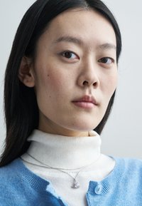 Ανοιχτό μπλε ζακέτα με στρογγυλή λαιμόκοψη, φορεμένη πάνω από ένα λευκό ζιβάγκο. Ένα ασημένιο αλυσίδα κολιέ διαθέτει ένα τριγωνικό μωβ παντατιφ.