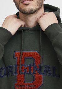 Khaki green hoodie featuring a prominent embroidered "B" in orange and blue, with a textured finish. Drawstrings are visible around the neck.