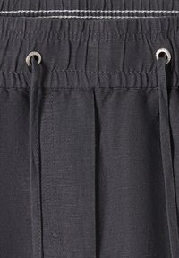 Gros plan sur un pantalon en tissu noir avec une ceinture élastique, des coutures blanches et des cordons noirs passant à travers des œillets métalliques.