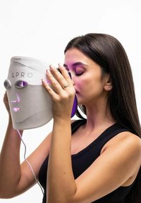 Vrouw die een wit LED-lichttherapemasker met het opschrift "STYLPRO" dicht bij haar gezicht houdt, met gesloten ogen en een zwart mouwloos topje aan.