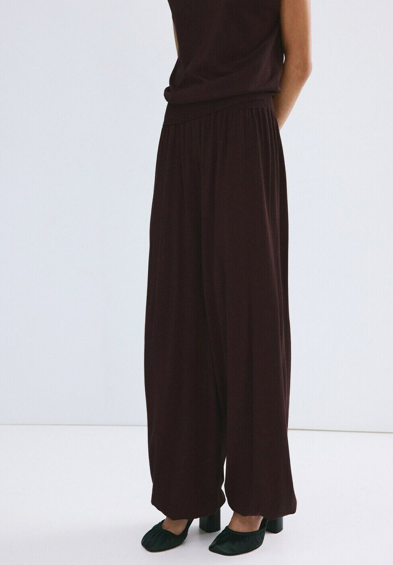 Personne portant un pantalon large marron foncé ample, associé à des chaussures à talons noires, debout devant un fond blanc uni.