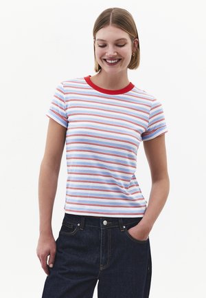 Lachende vrouw met kort haar draagt een wit gestreept t-shirt met korte mouwen en rode rand, en donkerblauwe spijkerbroek, één hand in de zak.