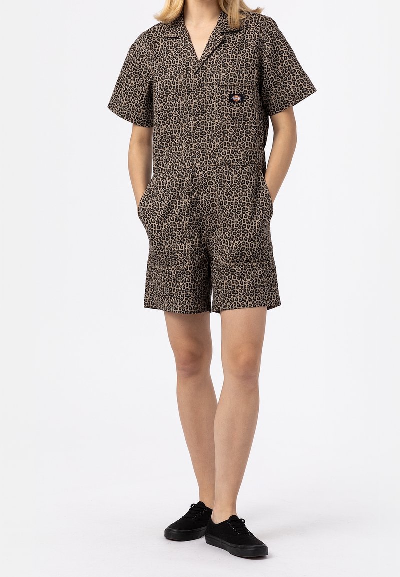 Kortjarige jumpsuit met luipaardprint in tan en zwart, met een kraag, knoopsluiting aan de voorkant en zijzakken. Gecombineerd met zwarte sneakers.