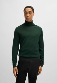 Donkergroene coltrui, met ribbenmanchetten en onderrand, aansluitend ontwerp, lichtgewicht breiwerk, gladde textuur, gedragen met zwarte broeken.