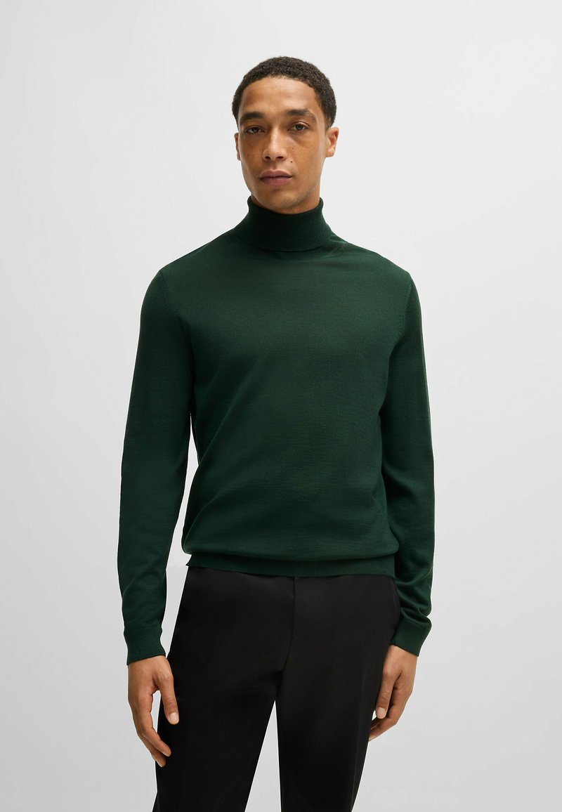 Donkergroene coltrui, met ribbenmanchetten en onderrand, aansluitend ontwerp, lichtgewicht breiwerk, gladde textuur, gedragen met zwarte broeken.