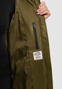 Olijfgroene jas met een glanzende afwerking, voorzien van een binnenzak met een zwarte ritssluiting en een label met de tekst "Een warme omhelzing van stof."