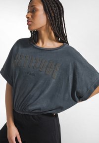 Camiseta oversize de color gris oscuro con cuello redondo, con estampado texturizado "ACTITUD". Mangas cortas y detalle de cintura fruncida.