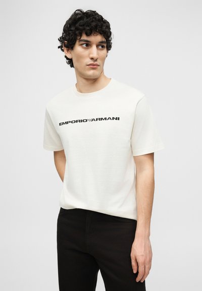 Άνδρας με σγουρά μαλλιά που φοράει λευκό t-shirt Emporio Armani και μαύρο παντελόνι, κοιτάζοντας πλάγια σε μονοχρωματικό γκρι φόντο.