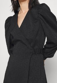 Svart omslagsklänning med puffärmar, texturerad tyg med ett subtilt mönster, insvängd midja med knapprad och djup V-ringning.