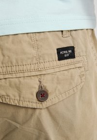 Shorts en tissu beige clair avec une poche plaquée ornée d'une étiquette noire "PETROL IND. & CO." et un bouton avec un accent décoratif rouge.
