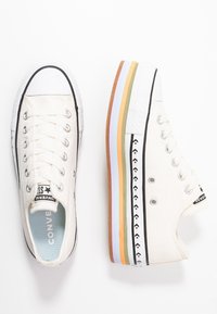 Vita canvas sneakers med en tjock, flerfärgad plattformssula. Har svarta detaljer, ögla och stjärnlogotyp på hälen.