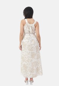 Frau mit langen dunklen Haaren, die ein ärmelloses, weißes Maxikleid mit beigen Blumenmustern trägt und barfuß vor einem weißen Hintergrund steht.